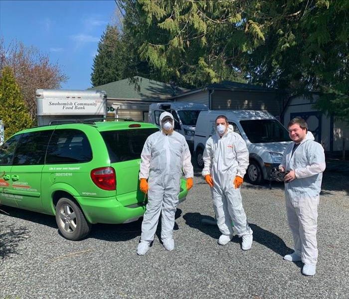 3 employees in Personal protective gear standing in a gravel lot next to a green SERVPRO van